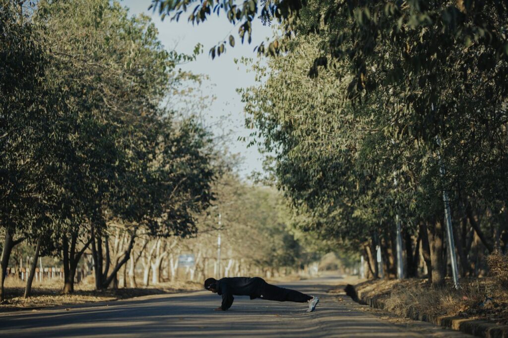 A man performs push-ups outdoors on a tree-lined pathway under the sun.